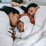 A mixed gender couple cuddles together with a baby in between them on a bed, sharing a white blanket.